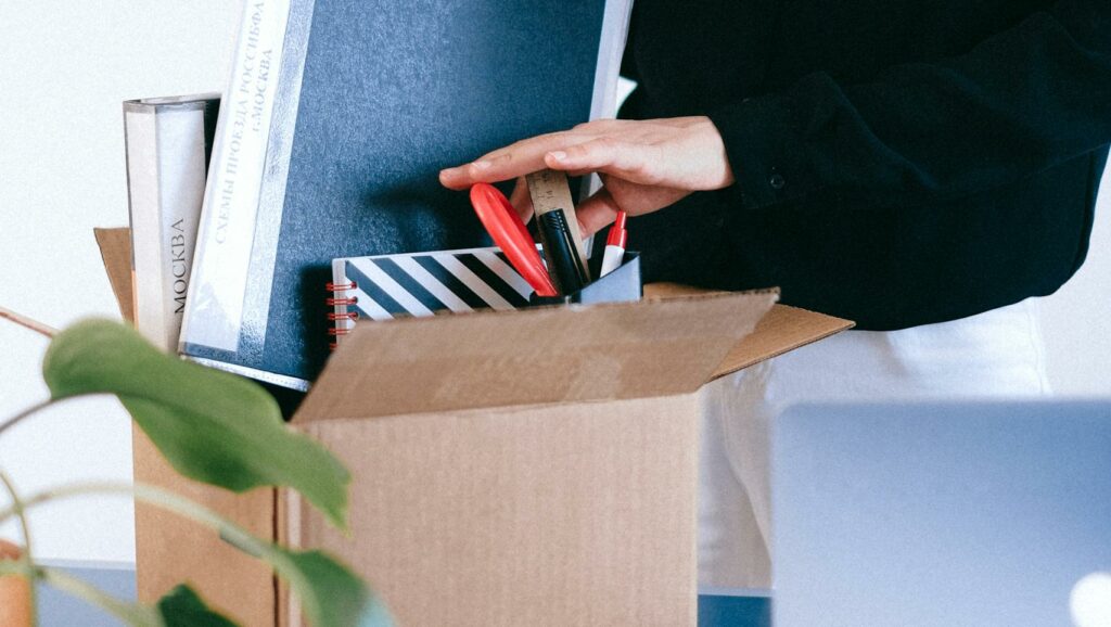 Adult woman organizing office supplies into a cardboard box, possibly symbolizing job change or relocation.